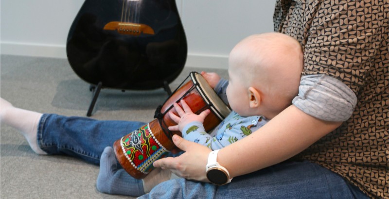 Baby sitter på mors fang og spiller på en tromme. Foto.