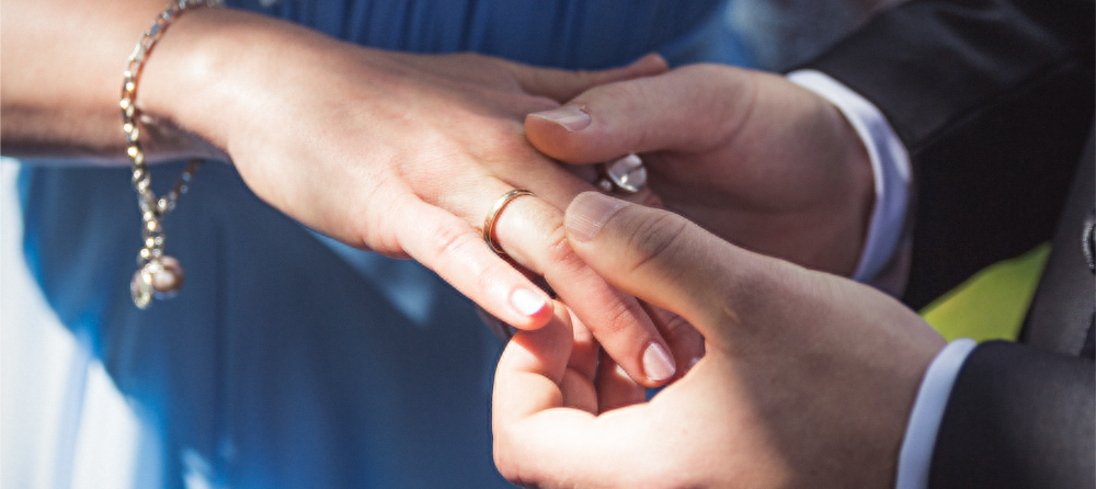 Nærbilde av en brud og brudgom under vielsen, der brudgommen setter en gullring på brudens finger. Bruden har en lyseblå kjole og et armbånd med en perle, mens brudgommen er kledd i en mørk dress. Sollyset skaper en varm og romantisk stemning.