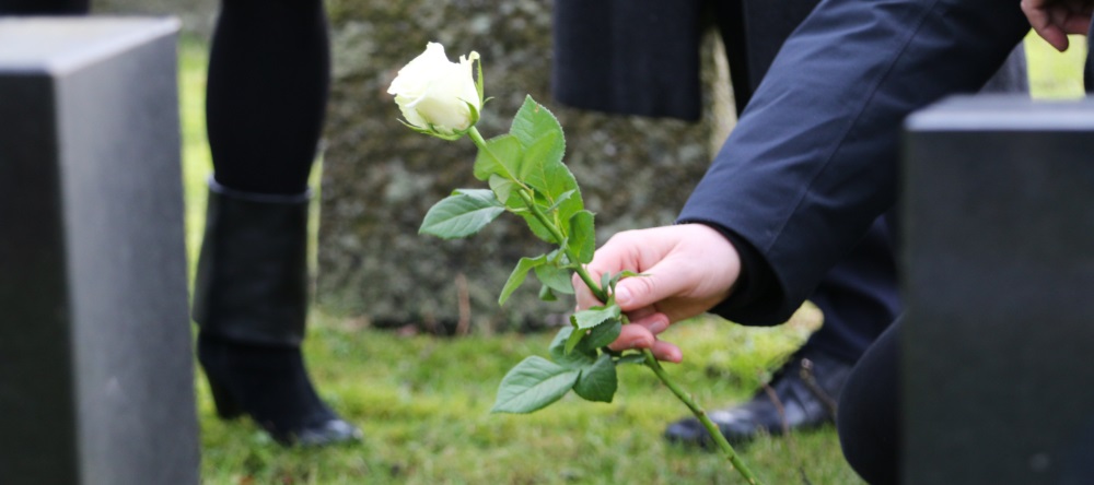 En person kledd i mørke klær legger en hvit rose på en grav. Bildet viser en nærbilde av hånden som holder blomsten, omgitt av gravsteiner og gress. I bakgrunnen står andre sørgende i svarte klær. Foto.